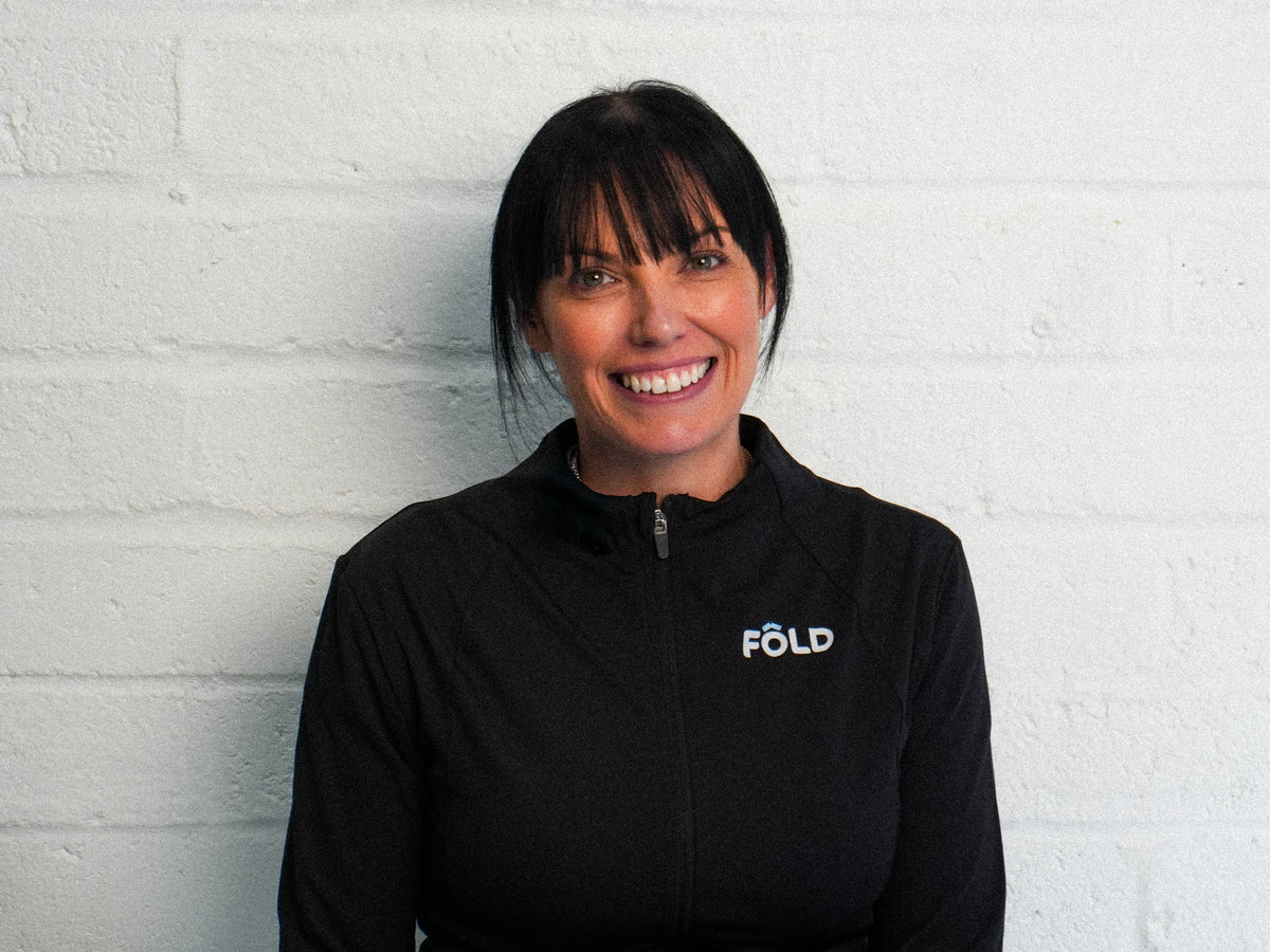 Smiling woman in a black jacket with FÖLD logo against a white brick wall, showcasing a friendly atmosphere.