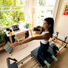 Woman using 3 Months App Membership on a Reformer bed in a bright, cozy home studio.