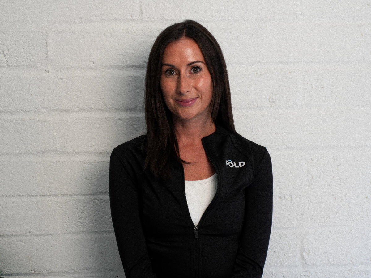 Smiling woman in a black zip-up jacket against a white brick wall, showcasing a casual and confident pose.
